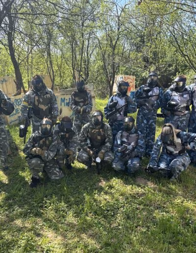 Grupa ljudi u maskirnim odijelima i zaštitnim kacigama pozira s paintball puškama na travnatom području okruženom drvećem.