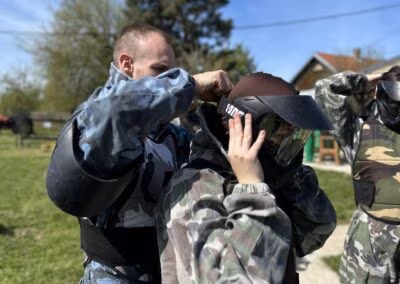 Osoba u maskirnoj uniformi pomaže drugoj osobi popraviti zaštitnu kacigu na paintball terenu, okruženi zelenim travnjakom i drvećem pod vedrim nebom.