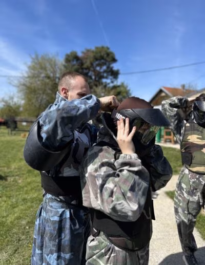 Osoba u maskirnoj uniformi pomaže drugoj osobi popraviti zaštitnu kacigu na paintball terenu, okruženi zelenim travnjakom i drvećem pod vedrim nebom.