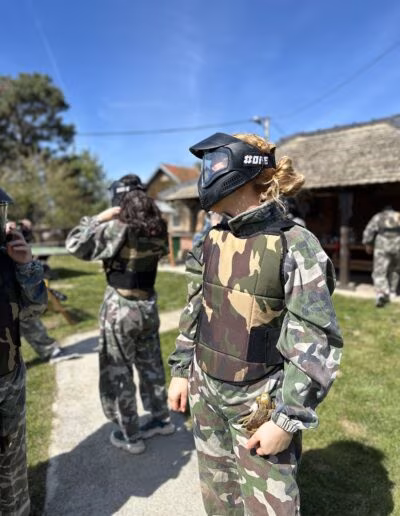 Osoba u maskirnoj odjeći i zaštitnoj kacigi stoji na otvorenom prostoru, vjerojatno tijekom paintball aktivnosti, s drvenom kućicom u pozadini.
