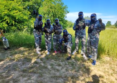 Skupina ljudi u maskirnim odijelima s opremom za paintball stoji na livadi okruženoj šumom, spremni za igru.