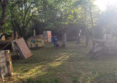 Prostor za paintball na otvorenom s drvenim preprekama i rekvizitima, okružen drvećem i travnjakom, uz sunčevu svjetlost koja prodire kroz krošnje.