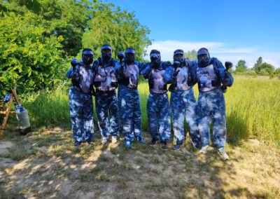Grupa ljudi u maskirnim uniformama i zaštitnim kacigama stoji na travnatom području, držeći paintball puške, okruženi zelenilom i drvećem ispod plavog neba.