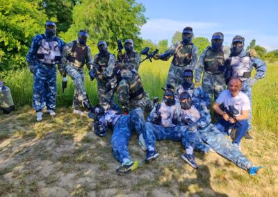 Skupina ljudi u maskirnim odijelima za paintball s kacigama i puškama, stoje i sjede na travnatom području, okruženi zelenilom, na sunčan dan.