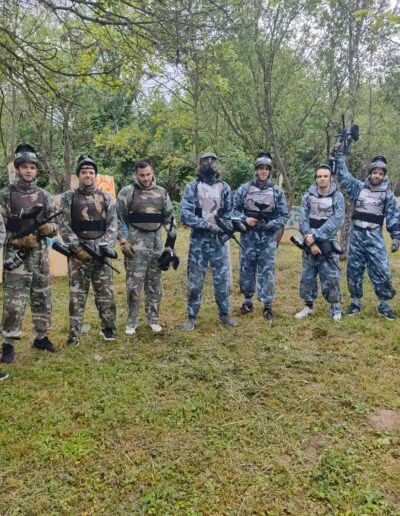 Skupina ljudi u maskirnim odijelima i zaštitnim kacigama drži paintball puške u prirodnom okruženju s drvenim preprekama.