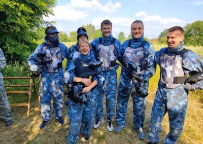 Grupa ljudi u plavim vojničkim uniformama za paintball stoji na otvorenom polju, noseći maske i držeći paintball puške, okruženi zelenilom i drvećem.