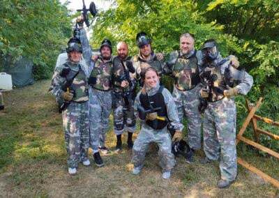 Grupa ljudi u maskirnim uniformama i zaštitnoj opremi drži paintball puške i pozira na otvorenom prostoru s drvećem u pozadini.
