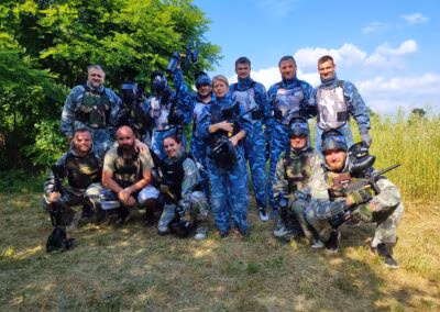 Skupina ljudi u paintball opremi pozira na otvorenom, neki nose maskirne uniforme, a drugi plave kamuflaže, držeći paintball puške, okruženi zelenilom i pod vedrim nebom.